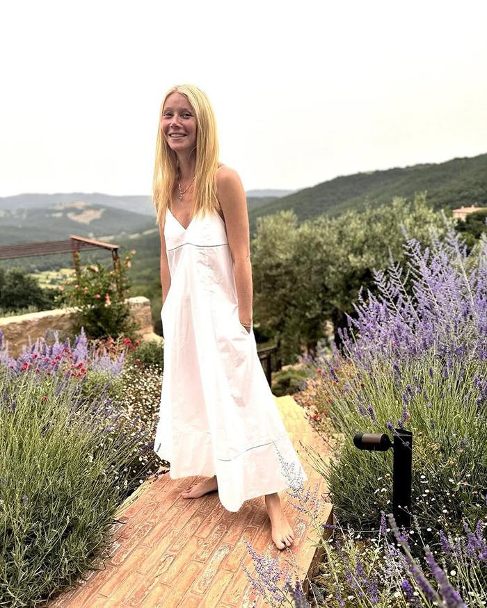 Gwyneth Paltrow standing barefoot on a garden path surrounded by flowers and greenery, smiling outdoors. Gwyneth Paltrow standing barefoot on a garden path surrounded by flowers and greenery, smiling outdoors.