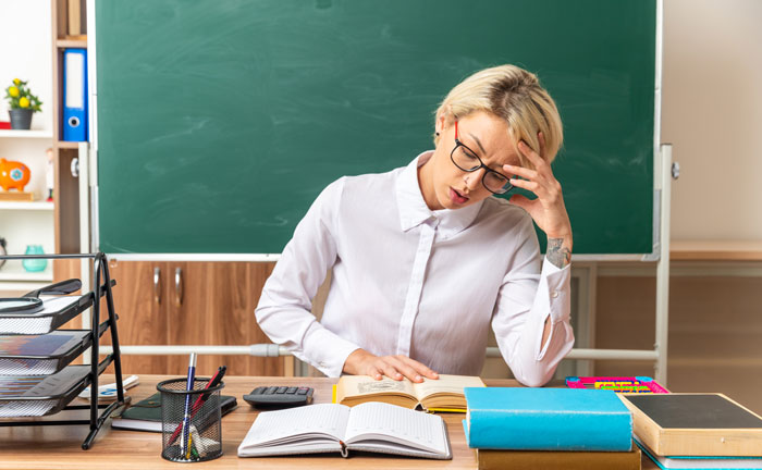 Teacher sitting at desk in classroom with books, appearing stressed while preparing for school students in hallways lanes.