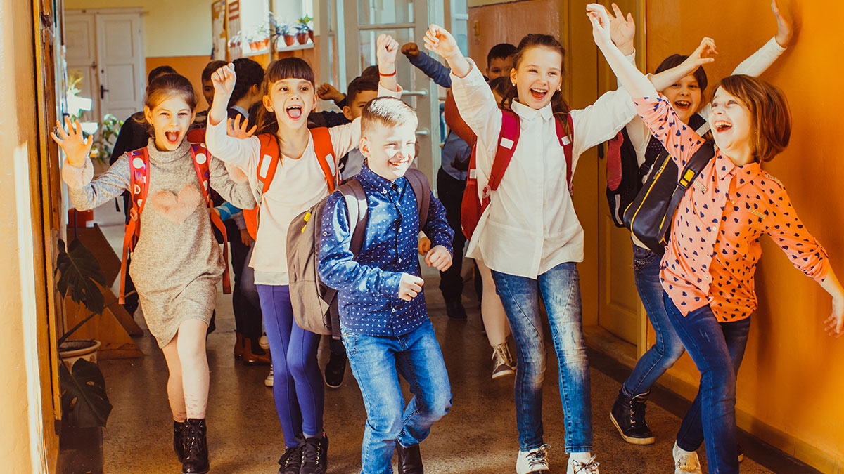 Students laughing and pretending to drive imaginary cars along school hallway lanes, showing playful ridicule.
