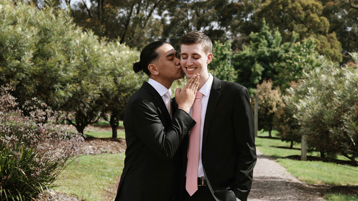 Gay couple dressed formally outdoors, sharing a tender moment with one partner kissing the other on the cheek.