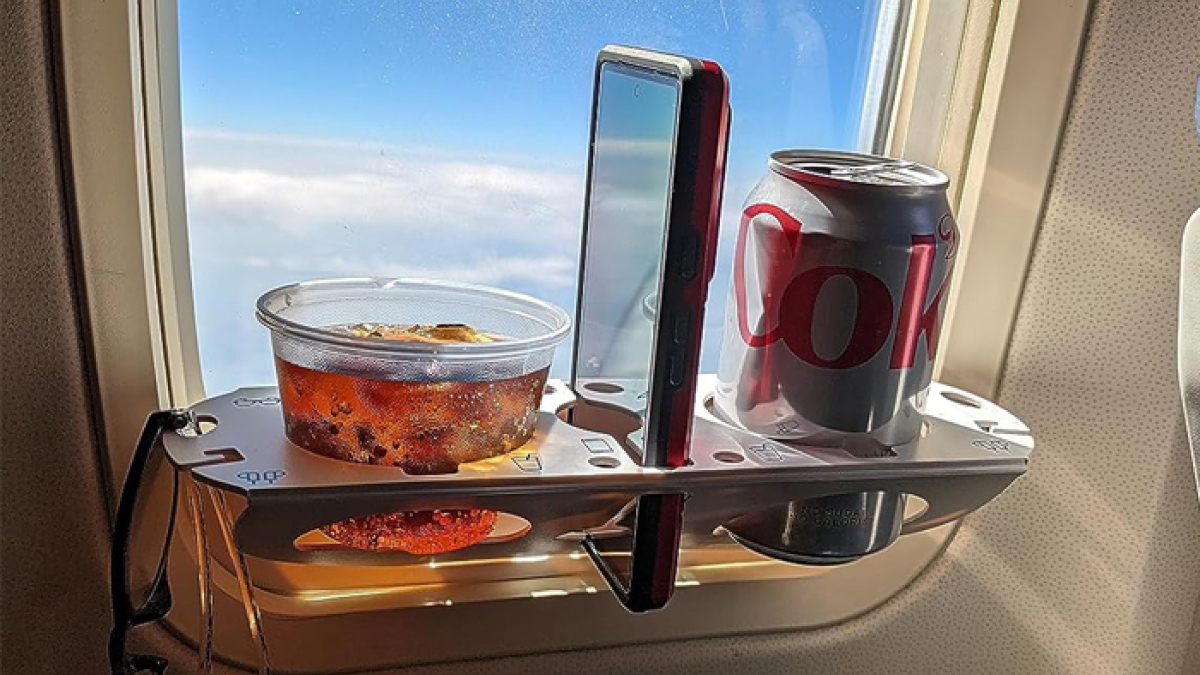 Airplane window tray holding a drink, a phone, and a can, showcasing travel essentials for a hot girl summer itinerary.
