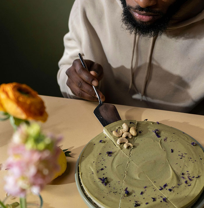 Man in beige hoodie cutting and serving a green cake slice topped with cashews on a beige table with flowers nearby. Man in beige hoodie cutting and serving a green cake slice topped with cashews on a beige table with flowers nearby.