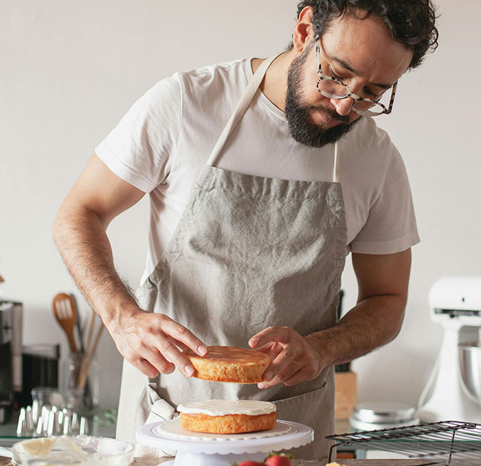 Man wearing glasses and apron carefully assembling a birthday cake in a kitchen, baking and preparing dessert. Man wearing glasses and apron carefully assembling a birthday cake in a kitchen, baking and preparing dessert.