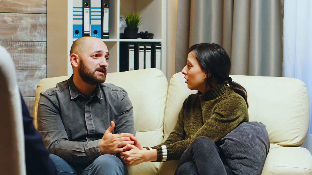 Husband and wife having a serious talk on couch, wife showing concern after husband's emotional confession of lingering feelings.