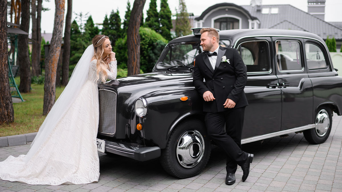 Bride and groom posing by a classic black car highlighting husbandu2019s car service business on wedding day.
