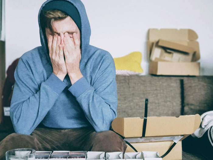 Man in hoodie covering face with hands, sitting amid unpacked boxes, illustrating travel difficulties and a weird quirk. Man in hoodie covering face with hands, sitting amid unpacked boxes, illustrating travel difficulties and a weird quirk.