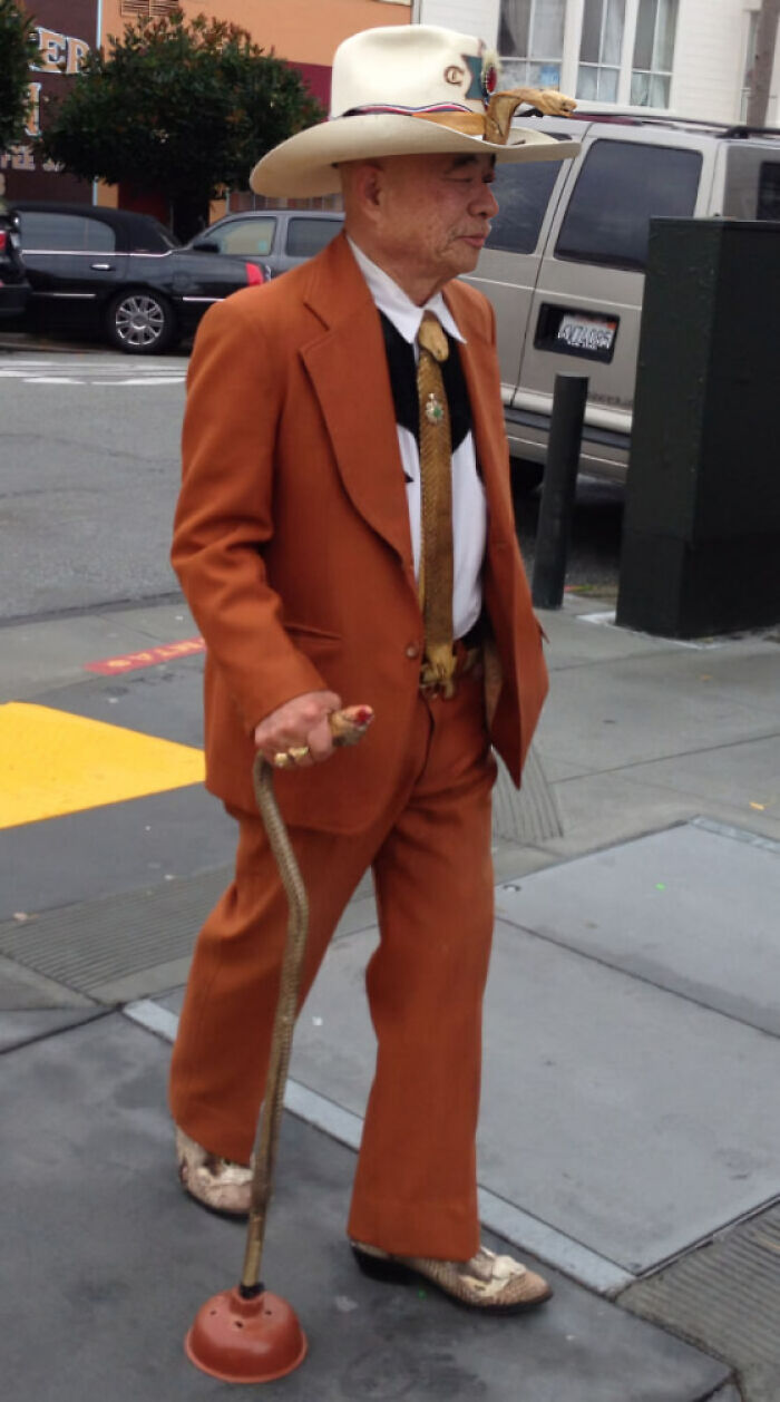 Elderly man in an orange suit walking confidently with a quirky plunger cane, showing suspiciously unbothered attitude.