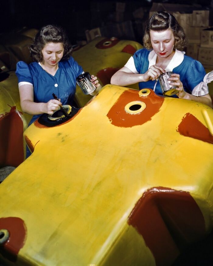 Two women in blue dresses painting a large yellow object, showcasing women at work during WWII in a factory setting.