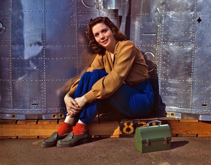 Woman worker during WWII sitting by aircraft metal panels with toolbox, representing women at work in the WWII era.