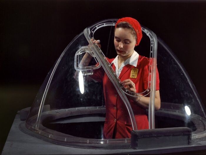 Woman at work during WWII assembling aircraft parts, wearing a red uniform and headscarf inside a clear cockpit frame.
