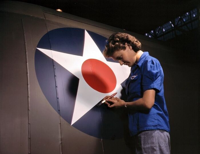 Woman in blue uniform painting a military aircraft emblem, representing women at work during WWII in a factory setting.