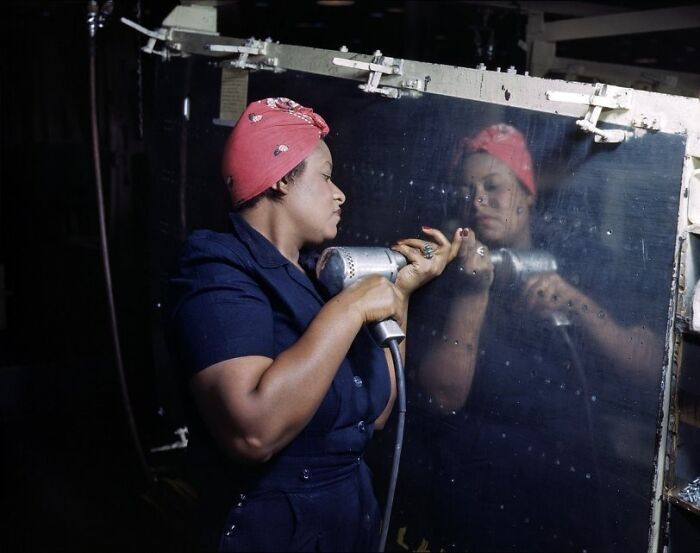 Woman at work during WWII wearing a red bandana and using a power tool on a metal surface, showcasing women at work.