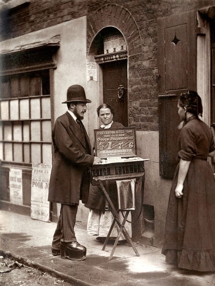 Man in 1800s London selling cough lozenges on street while women in period clothing observe, showing poorest living conditions.
