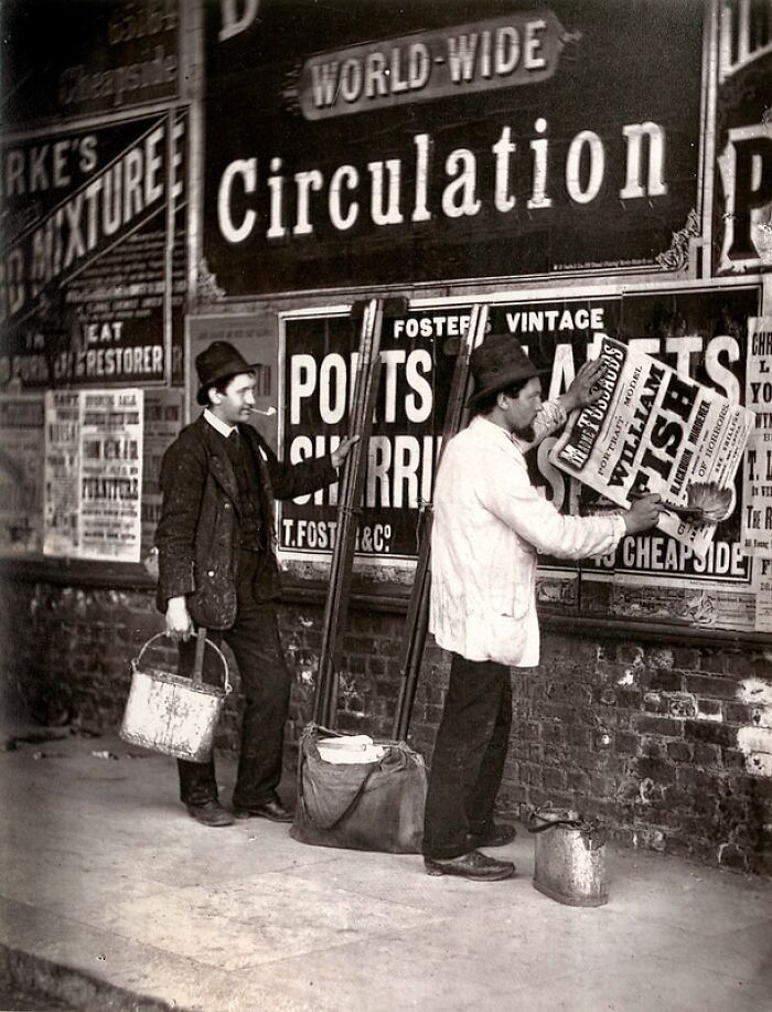 Two men posting vintage advertisements on a brick wall, depicting life of London's poorest in the 1800s.