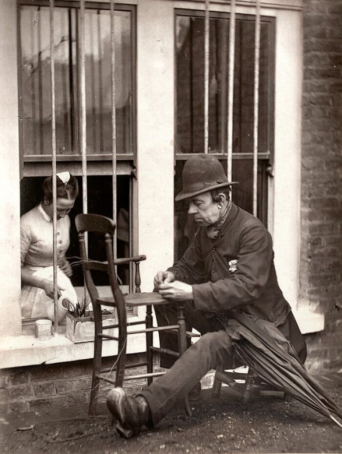 Man sitting outside barred window repairing chair while woman inside tends plants, showing how London's poorest lived in 1800s.