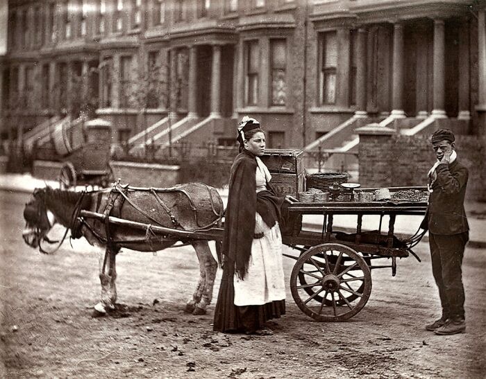 London's poorest woman selling goods from a horse-drawn cart on a 1800s urban street with brick houses in the background