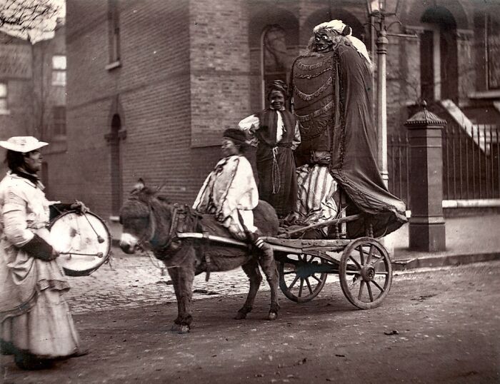 Victorian street scene with London’s poorest riding a donkey cart and a woman playing a drum in 1800s London.