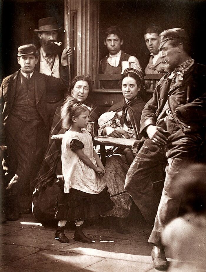 Group of London’s poorest people in the 1800s, including women, children, and men in worn clothing inside a small space.