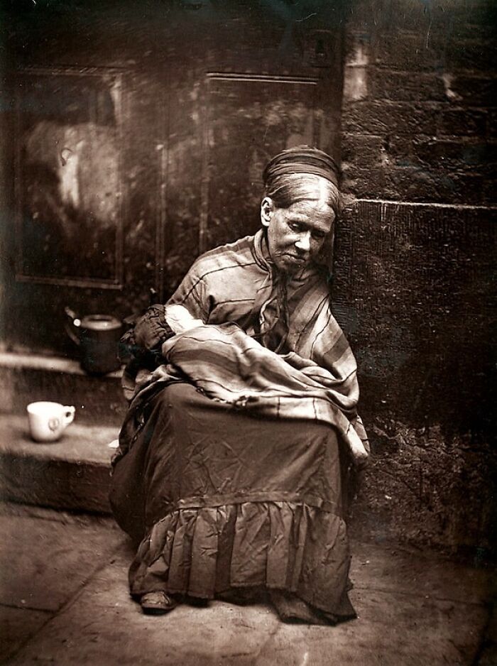 Elderly woman in 1800s London, wrapped in worn clothing, sitting against a wall showing poorest living conditions.