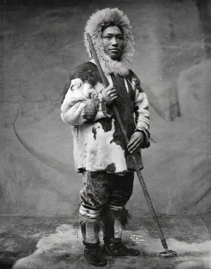 Historic black and white photo of an Inuit man in traditional fur clothing holding a spear, capturing Inuit spirit.