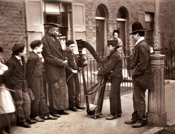 Group of London’s poorest children and a man performing street music in the 1800s Victorian neighborhood street scene.