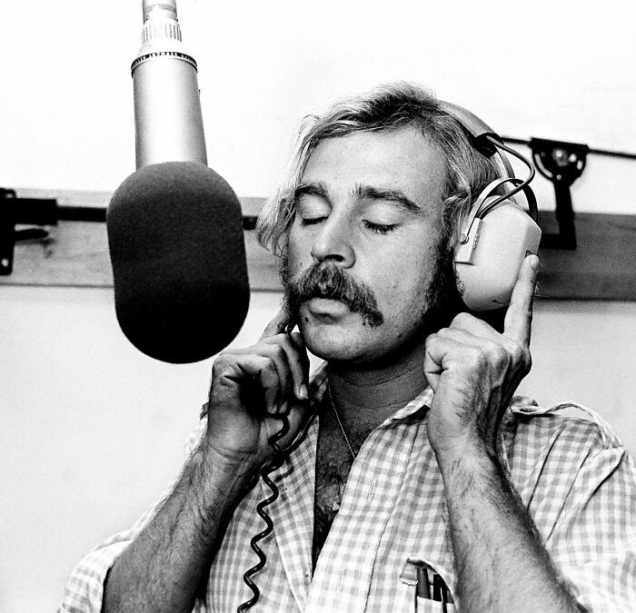 Black and white photo of 1970s rock legend wearing headphones and singing into a studio microphone during recording session