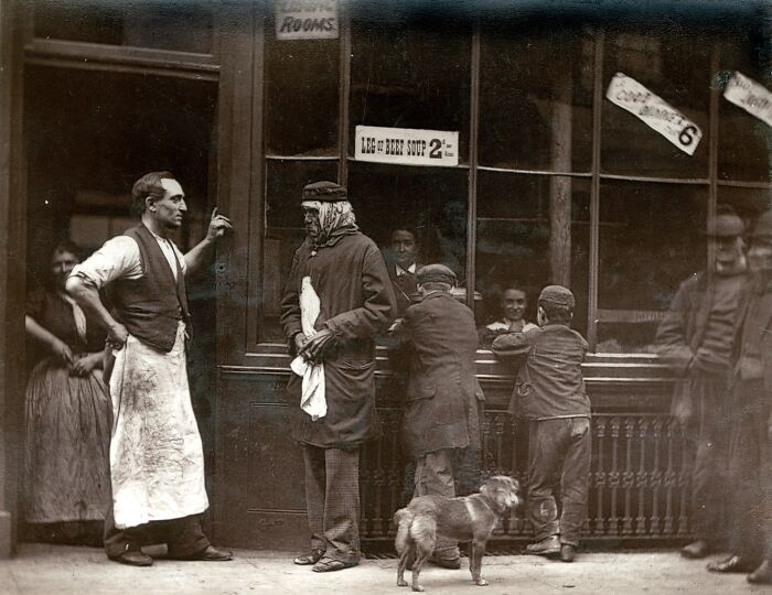 London's poorest in the 1800s outside a soup kitchen with people and a dog on a street scene.