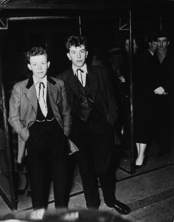 Two young men dressed in classic 20th century Teddy Boy fashion leaning against a metal railing at night.