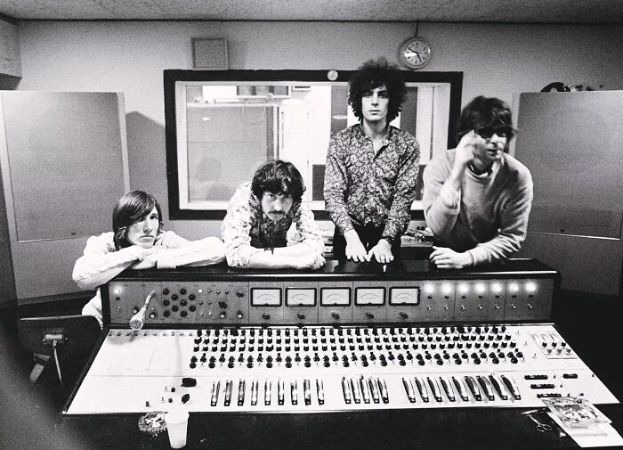 1970s rock legends posing behind vintage studio mixing console during a music recording session in black and white.