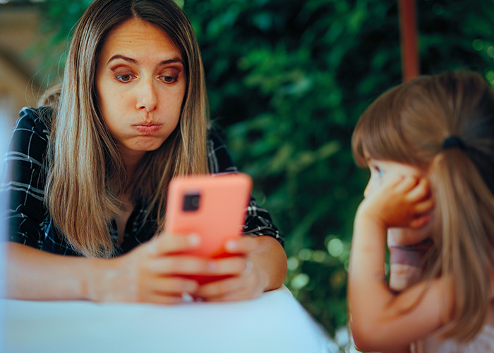 Momfluencer making exaggerated face at phone while daughter looks on, capturing staged moments for online fame controversy Momfluencer making exaggerated face at phone while daughter looks on, capturing staged moments for online fame controversy