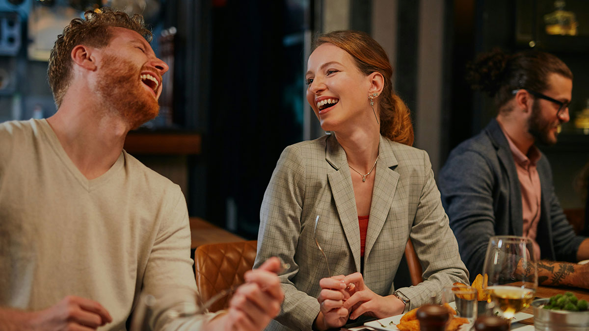 Three friends enjoying a lively dinner while sharing laughs, illustrating inlaws day and heritage information discussion.