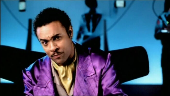 Man in a shiny purple suit with a yellow shirt, sitting indoors with blue lighting and abstract decorations behind.