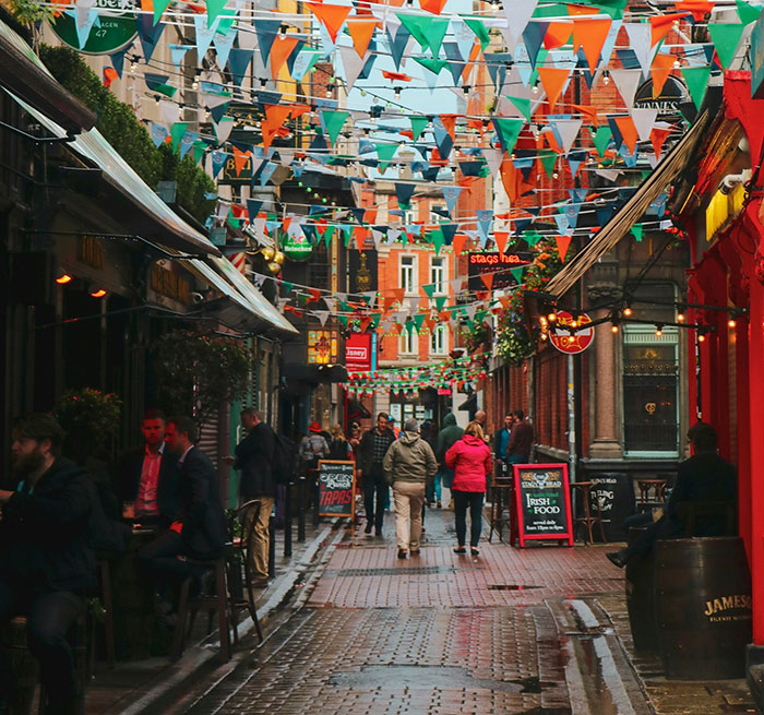Crowded narrow Irish street with colorful flags overhead and people walking near pubs and outdoor seating areas. Crowded narrow Irish street with colorful flags overhead and people walking near pubs and outdoor seating areas.