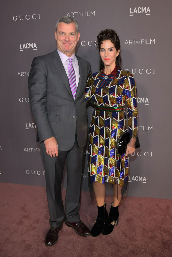 Jami Gertz at a formal event wearing a colorful geometric dress, standing next to a man in a gray suit and purple tie. Jami Gertz at a formal event wearing a colorful geometric dress, standing next to a man in a gray suit and purple tie.