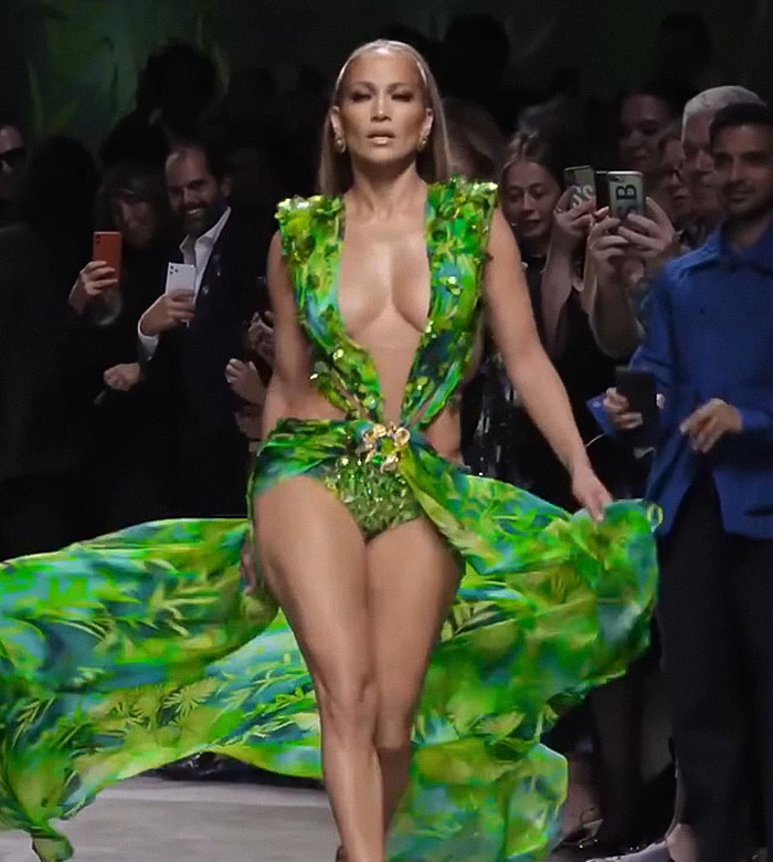 Jennifer Lopez walking runway in a green flowing dress, facing forward during a raunchy show with crowd capturing the moment. Jennifer Lopez walking runway in a green flowing dress, facing forward during a raunchy show with crowd capturing the moment.