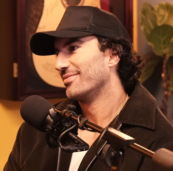 Man wearing a black cap and jacket speaking into a microphone during a casual podcast or interview session. Man wearing a black cap and jacket speaking into a microphone during a casual podcast or interview session.