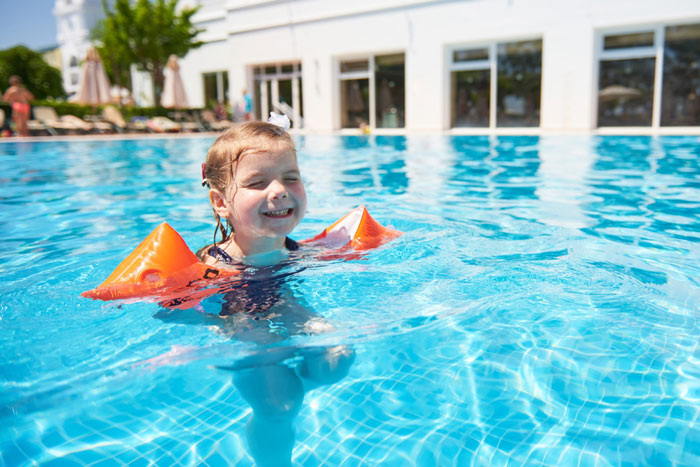 Child wearing orange floaties happily swimming in a hotel pool on a sunny day near hotel building and lounge chairs.
