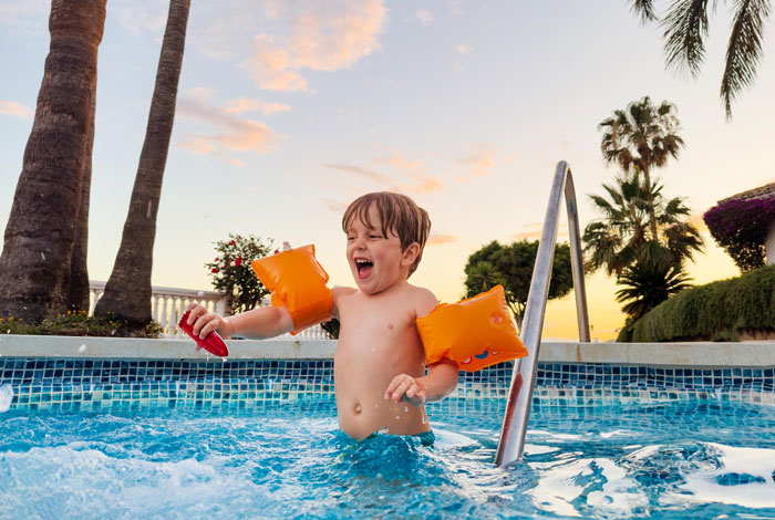 Young boy wearing floaties playing joyfully in a hotel pool at sunset near palm trees and tropical plants.