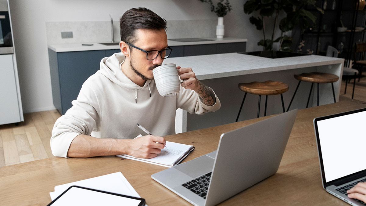 Man working on laptop from home, taking notes and drinking coffee, symbolizing person gets laid off and lands better job.