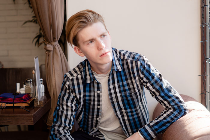 Teen sitting indoors, wearing a plaid shirt, looking thoughtful while coping with loss and working multiple jobs. Teen sitting indoors, wearing a plaid shirt, looking thoughtful while coping with loss and working multiple jobs.