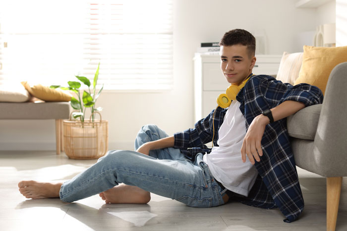 Teen sitting on floor near couch with headphones, representing a hardworking teen facing family challenges and survival struggles. Teen sitting on floor near couch with headphones, representing a hardworking teen facing family challenges and survival struggles.