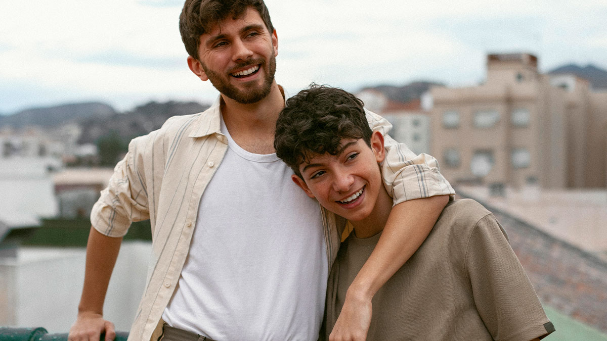 Teen with curly hair leaning on older smiling man outdoors, representing teen who works two jobs to survive.