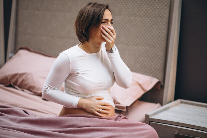 Pregnant woman sitting in bed holding her belly with a hand over her mouth, reflecting shock and distress about father and roomie.