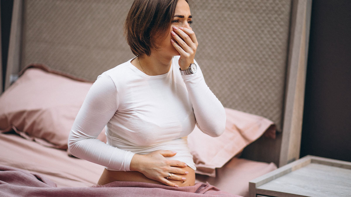 Pregnant woman sitting on bed, holding belly and covering mouth, expressing shock and concern about her situation.