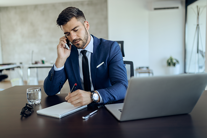 Man in a blue suit making a phone call while taking notes at a desk with a laptop and glass of water in a modern office.