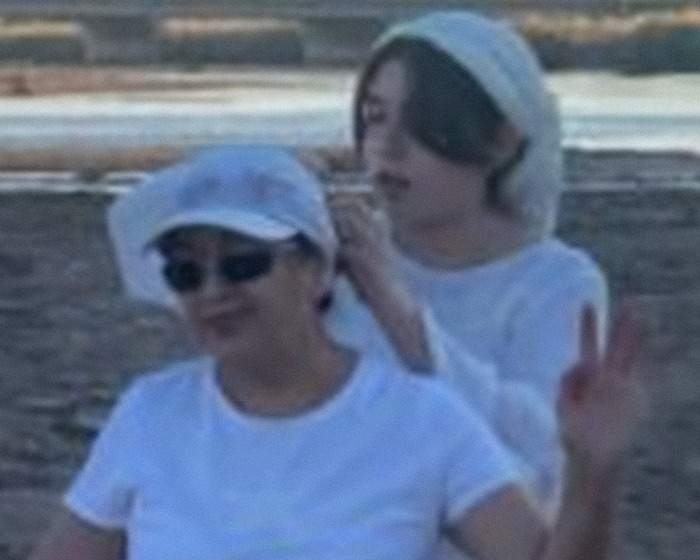 Two women outdoors near water, one fixing the other's headscarf, with calm expressions in a casual setting. Two women outdoors near water, one fixing the other's headscarf, with calm expressions in a casual setting.