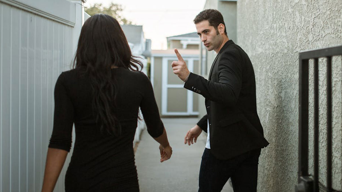 Man gesturing with finger while walking and talking to a woman in a narrow outdoor walkway setting