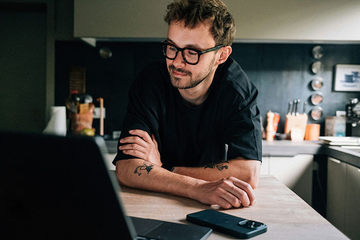 Man with glasses and tattoos sitting at kitchen table looking at laptop, illustrating father and son lock horns conflict. Man with glasses and tattoos sitting at kitchen table looking at laptop, illustrating father and son lock horns conflict.