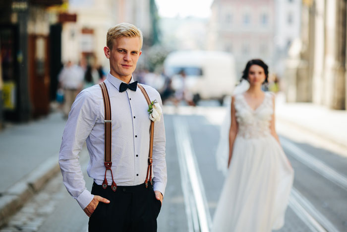 Groom standing on city street in suspenders and bow tie while bride in white wedding dress walks behind him. Groom standing on city street in suspenders and bow tie while bride in white wedding dress walks behind him.