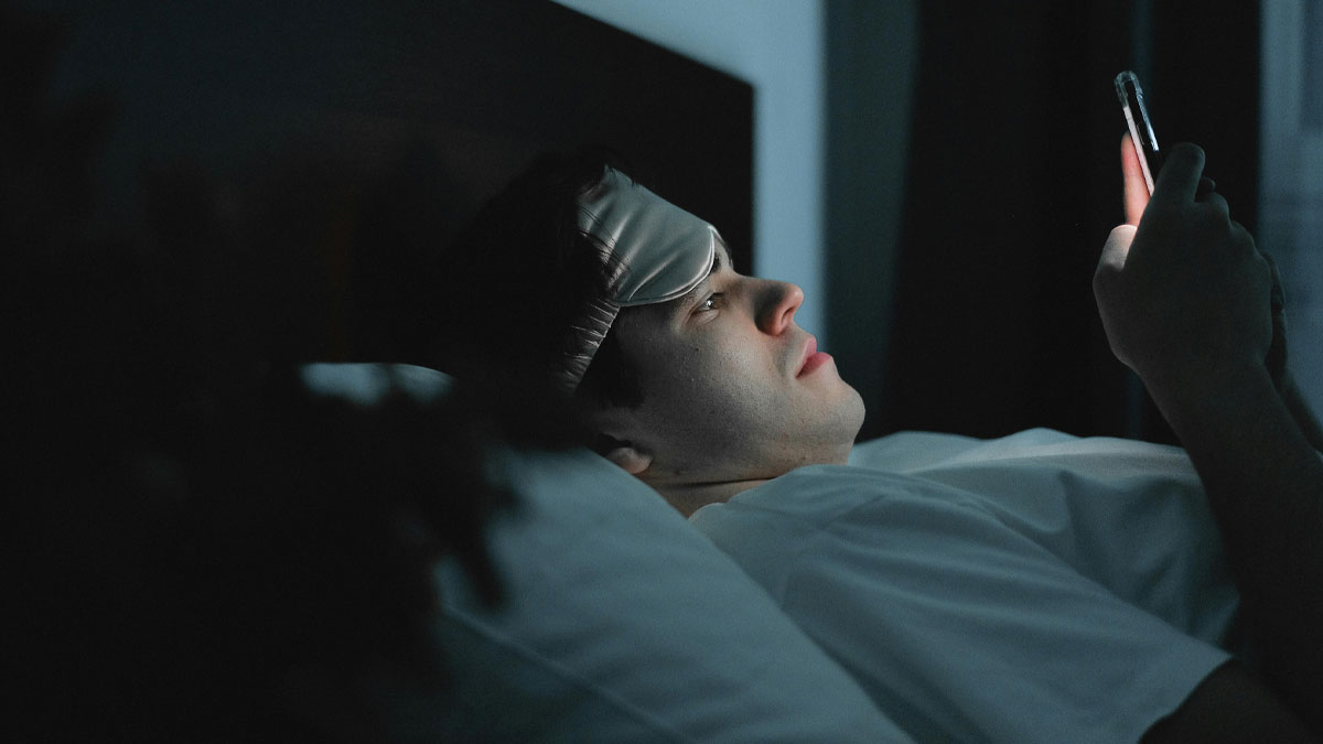 Man lying in bed at night using smartphone with sleep mask on forehead, illustrating micro cheating concept.